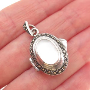 May include: Oval silver pendant with a white center and marcasite accents. The pendant has a hinged design and a small loop for a chain. The pendant is held in a hand.