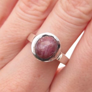 May include: A silver ring featuring a round, cabochon-cut, deep pinkish-red gemstone set in a smooth, polished metal bezel. The band is plain and metallic, showcasing a simple design that highlights the natural texture and color of the stone.