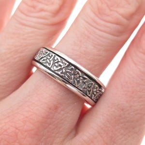 May include: A silver-toned metal ring featuring a continuous band with an embossed, repeating intricate knotwork pattern. The design consists of interconnected loops and lines that create a textured, recessed appearance across the surface.