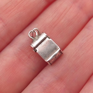 May include: A small, rectangular silver charm featuring a smooth, flat front surface and a rounded top section with an attached circular jump ring. The metal has a polished finish with subtle signs of wear and oxidation in the recessed areas.