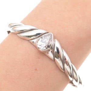 May include: A polished silver cuff bracelet featuring a twisted rope-like design and a single pear-shaped clear gemstone set in the center. The metal has a high-shine finish, and the band curves around the wrist with a smooth, continuous structure.