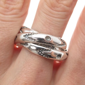May include: A silver-toned ring with an interwoven design. The ring features a textured surface with small, decorative details and circular cutouts. The ring is worn on a finger, showcasing its intricate craftsmanship and metallic sheen.