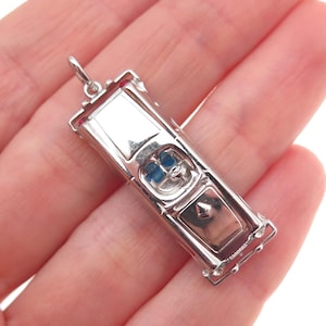 May include: A silver-toned pendant in the shape of a classic car. The pendant has a detailed design with a rectangular body, a windshield, and a small loop for a chain. The car's interior is blue. The pendant is held in a hand.