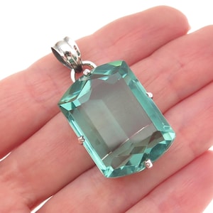 May include: A rectangular, faceted, light green gemstone pendant set in a silver-toned metal frame with four small prongs at the corners and a polished metal bail at the top for attachment to a chain or cord, showcasing a clear, glassy texture.