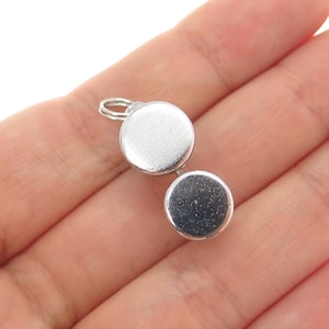May include: A small, silver-colored pendant with two circular, hinged compartments. The pendant has a loop at the top for attaching to a chain. The surface of the pendant is smooth and reflective, with a slight shine.