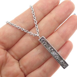 May include: A silver-toned pendant necklace featuring a rectangular pendant with an ornate, scrolling design. The pendant is suspended from a delicate chain. The necklace is held in a hand, showcasing the intricate details of the jewelry.