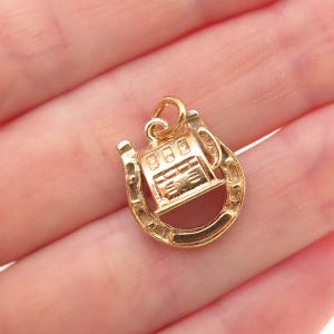 May include: A gold-colored charm featuring a slot machine design set within a horseshoe. The charm has a loop at the top for attachment. The slot machine has three visible reels. The horseshoe is a classic U-shape.