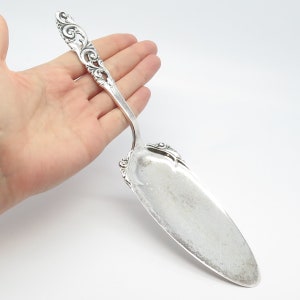 May include: A silver cake server with an ornate handle and a wide, curved blade. The handle features detailed scrollwork and flourishes. The server is designed for serving cakes and pastries.