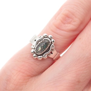 May include: A silver ring featuring an oval abalone shell center stone. The ring has a decorative, beaded border and a split shank design. The abalone shell displays iridescent colors of green, blue, and purple. The ring is shown on a finger.