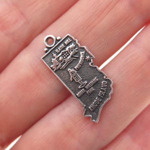 May include: A silver-toned metal charm shaped like the state of Rhode Island. The surface features embossed text reading Slater Mill, Providence, Roger Williams Park, and Rhode Island, along with small detailed illustrations of these landmarks.