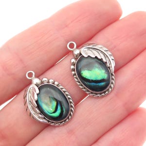 May include: Two oval pendants featuring iridescent abalone shell cabochons set in silver-toned metal. Each pendant is framed by a twisted rope border and accented with a sculpted leaf design on one side, topped with a small circular jump ring.