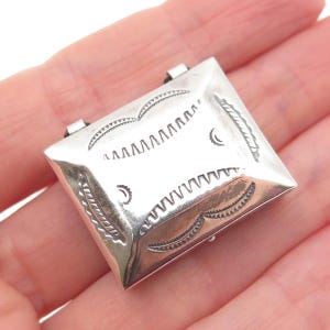 May include: A rectangular silver box with a hinged lid, featuring stamped geometric patterns including curved lines and small serrated markings on the top surface. The metal has a polished, reflective finish with visible hinge components.