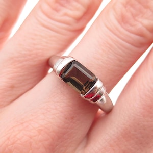 May include: A silver ring featuring a rectangular, smoky brown gemstone. The ring has a simple band with decorative silver accents on either side of the stone. The gemstone is set in a bezel setting, and the ring is shown on a finger.