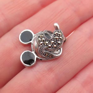 May include: A silver-toned charm in the shape of a mouse's head, featuring black enamel ears and a face adorned with small, sparkling stones. The charm has a detailed, textured design.