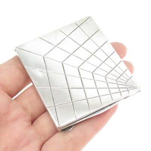 May include: A silver-colored, geometric compact mirror with a grid pattern. The square-shaped mirror has a hinged closure and is held in a hand against a white background. The grid design creates a 3D effect.
