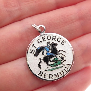 May include: A round silver pendant with the words "ST GEORGE BERMUDA" encircling a colorful image of a knight on horseback slaying a dragon. The knight wears blue and black, and the dragon is green.