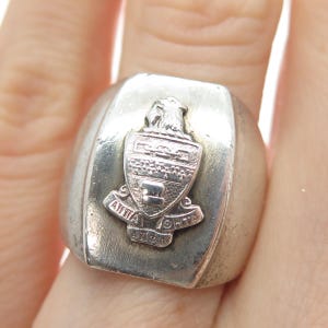 May include: A silver ring with a rectangular face. The ring features a raised emblem with a shield design, an eagle, and Greek letters. The ring is worn on a finger.