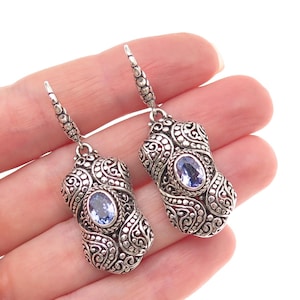 May include: A pair of silver-toned drop earrings. Each earring features an oval, light blue gemstone set within an ornate, filigree design. The earrings are suspended from a decorative hook.