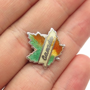 May include: A silver-toned maple leaf pin with green, orange, and yellow enamel detailing. The word "Canada" is written vertically in a cream-colored rectangle across the center of the leaf.