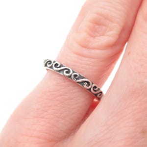 May include: A silver ring with a continuous, swirling scroll design. The ring is worn on a finger, showcasing the intricate pattern. The ring's design is a repeating pattern of elegant curves.