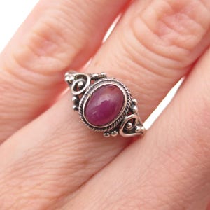 May include: A silver ring featuring an oval-shaped, deep pink cabochon gemstone set in a twisted metal border. The band is decorated with intricate silver scrollwork and small metal spheres, creating a detailed frame around the central stone.