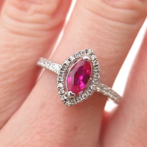May include: A silver ring featuring a marquise-cut, vibrant pink gemstone surrounded by a halo of small, clear stones. The ring's band is also adorned with small, clear stones.