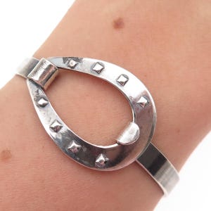 May include: A silver-toned bracelet with a horseshoe-shaped centerpiece. The bracelet features a series of square-shaped embellishments around the horseshoe. The bracelet is worn on a wrist.