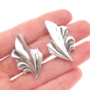 May include: A pair of silver-toned leaf-shaped earrings. Each earring has a curved, pointed design with intricate detailing, reflecting light. The earrings are displayed on a light background.