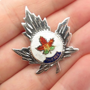 May include: Silver tone maple leaf pin with enamel detailing. The center of the pin features a red, orange, and green maple leaf with a white background. The word "QUEBEC" is written in blue around the maple leaf.