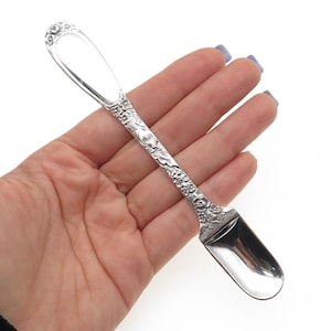 May include: A silver-toned metal spoon with a short, wide, polished bowl and a long, slender handle. The handle features an intricate, raised floral pattern that extends from the base of the bowl to the rounded, decorative tip of the spoon.