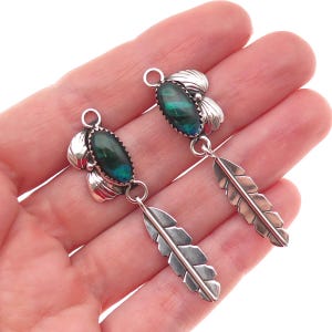 May include: A pair of silver earrings featuring oval-shaped dark green iridescent stones set in serrated bezels. Each earring includes a silver leaf-shaped charm dangling from the base, with a small circular loop at the top for attachment.