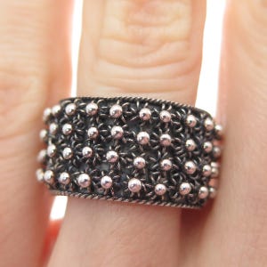 May include: A wide, silver-toned ring with a detailed, openwork design. The ring features a pattern of small, raised spheres connected by delicate, woven metal. The edges are finished with a twisted rope-like border, creating a textured and ornate appearance.