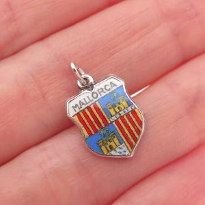 May include: A silver-toned Mallorca shield charm with a white border. The shield features a design with red and yellow stripes, a blue section with a castle, and the word "MALLORCA" at the top. The charm is held in a hand.