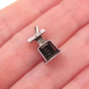 May include: A small silver-toned metal charm shaped like a vintage kitchen scale. The charm features a flat top platform, a central vertical support column, and a base with a visible internal coiled spring mechanism. It includes a small jump ring.