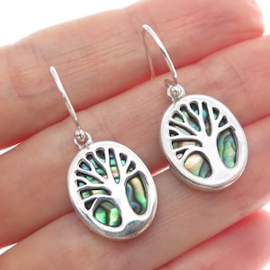 May include: A pair of silver-toned drop earrings. Each earring features an oval pendant with a tree of life design. The tree's interior is filled with iridescent abalone shell, displaying shades of green, blue, and purple. The earrings have a simple hook closure.