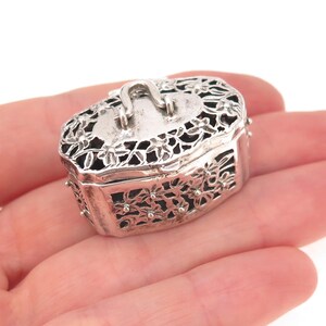 May include: A small, oval-shaped silver box featuring an intricate, openwork floral pattern across its lid and sides. The lid includes a small, arched handle at the center, and the entire piece has a polished, metallic finish with dark accents.