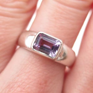 May include: A silver ring with a rectangular, purple gemstone. The gemstone is set in a bezel setting, and the ring has a smooth, polished band. The ring is shown on a finger.