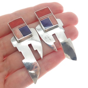 May include: A pair of silver earrings with a geometric design. Each earring features a rectangular top with red, pink, and blue inlays, and a stylized, elongated silver shape. The earrings are displayed against a white background.