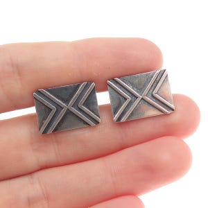 May include: A pair of silver-toned cufflinks, each square-shaped with a raised envelope design. The design features a central X and angled lines, creating a textured appearance. The cufflinks are held in a hand against a white background.