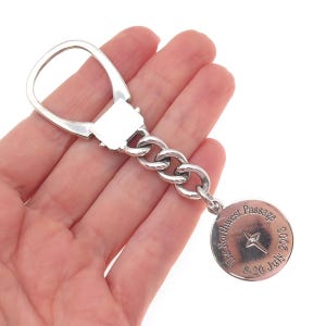 May include: A silver-toned metal keychain featuring a large oval ring, a short curb chain, and a circular pendant. The pendant is engraved with the text The Northwest Passage 8-20 July 2003 and features a small four-pointed star design in the center.