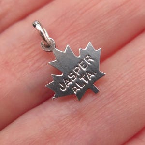 May include: A small, silver-colored charm in the shape of a maple leaf. The charm has the words "JASPER ALTA" engraved on it. The charm has a small loop at the top for attaching to a necklace or bracelet.