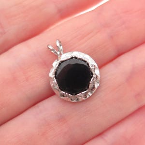 May include: A circular silver pendant featuring a smooth, polished black stone at its center. The silver frame has a textured, scalloped edge with a decorative pattern. A small silver bail is attached to the top for hanging on a chain.