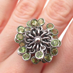 May include: A silver ring featuring a floral design with multiple round, light green gemstones. The ring's setting has a detailed, interconnected pattern, and the gemstones are evenly spaced around the central design.