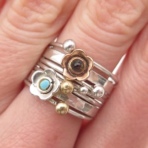 May include: A collection of silver rings, some with flower designs. One ring features a turquoise stone, while another has a dark stone center. Additional rings have gold and silver ball accents. The rings are stacked on a finger.