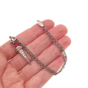 May include: A silver-toned pendant necklace featuring a dagger-shaped charm. The charm is adorned with clear crystals and a red gemstone. The necklace has a delicate chain design.