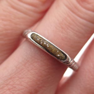 May include: A silver ring with a rectangular, light brown inlay. The ring has a textured, ridged design on the band. The inlay appears to have a speckled pattern. The ring is shown on a finger.