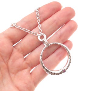 May include: A silver-toned necklace featuring a large, circular pendant with a hammered, textured surface. The pendant is suspended from a thick, round link chain and includes a single, small, square-set pink gemstone embedded in the metal rim.