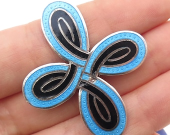 Puede incluir: Broche plateado con un diseño de cuatro pétalos. Cada pétalo presenta una forma negra curvada delineada en esmalte azul claro con un borde punteado. El broche se sostiene en una mano.
