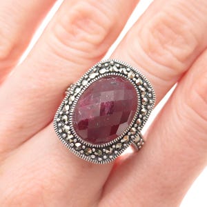 May include: A silver ring featuring a large, oval-shaped, faceted deep red gemstone at the center. The stone is surrounded by a decorative border of small, dark, metallic marcasite stones set in a textured, oxidized silver frame with a beaded edge.