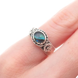 May include: A silver ring featuring an oval-shaped, iridescent gemstone. The ring has a floral design on the band and around the gemstone setting. The gemstone displays shades of blue, green, and teal.
